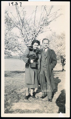Photograph of Rose and Milton Friedman in overcoats, standing in front of blossoming tree and body of water