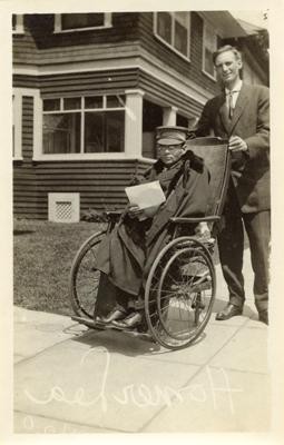 Photograph of Homer Lea, looking at the camera, being pushed in a wheelchair by an unidentified man