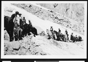 A group of people horseback riding along a snow-capped rocky mountainside, ca.1930