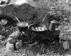 Bacon in a frying pan and other cooking items over a camp fire, California, ca.1910