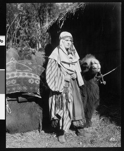 Man in Middle Eastern costume standing next to camel, ca. 1920