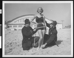 Two men in police costumes measuring a woman in a bathing suit, Venice Beach, ca.1920