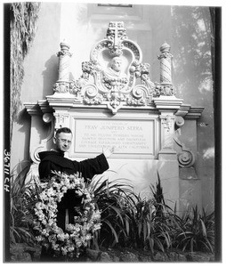 Friar gesturing to a memorial for Father Junipero Serra