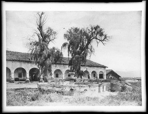 Fountain and two tall trees in front of Mission San Fernando, California, ca.1882