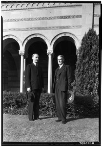 Two men in suits in front of a building with decorative arches