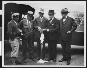 Five men looking at a map near a plane, showing one man in informal attire, ca.1930