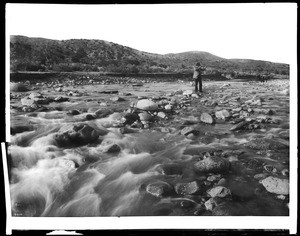 Man with rifle stands in mountain stream as it floods, east of Palmdale, California, ca.1920