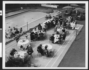 Diners at tables on a volleyball court near a swimming pool on what appears to be the grounds of a hotel, ca.1930