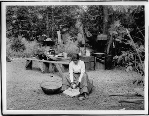 Paiute indian woman sitting on a small wooden crate grinding acorns on a matate stone in Yosemite, ca.1900