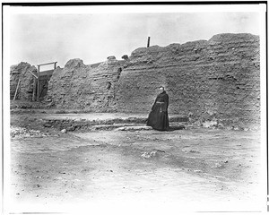 Friar standing in the ruins of the hall of Mission San Miguel's older church, ca.1900