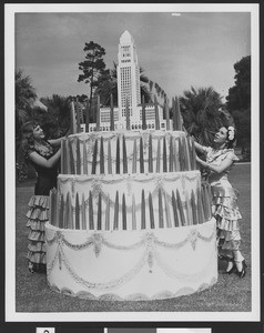 Two young women in Spanish costumes placing candles on a 5-ft tall cake that has a model of the Los Angeles City Hall on top, ca.1930