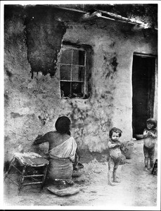 Hopi Indian woman whitewashing her house while two children play nearby, ca.1900