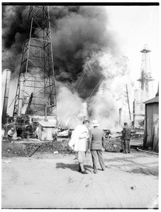 Oil fire at an unidentified oil field, showing three men watching from a distance