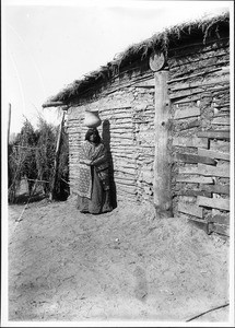 Yuma Indian woman carrying an "olla" on her head outside a building, ca.1900