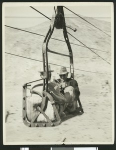 Blurred view of two men at a salt mine sitting on a cable that appears to be in motion, ca.1910