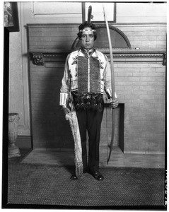 Portrait of Wilbur Anton with Indian ceremonial robes of beads and a bow and arrows at the Wigwam Club, October 4, 1928