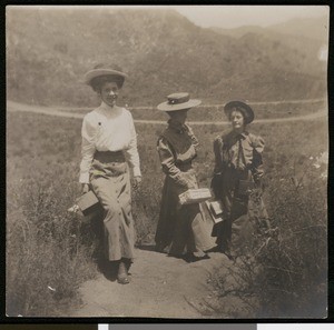 Three women with picnic lunches(?) and cameras coming up a hillside trail in the Cahuenga pass, 1900-1909