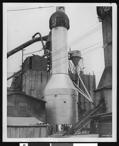 Exterior of an unidentified factory, showing a group of people gathered near the base of a smokestack, ca.1930