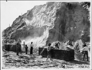 Group of thirteen miners working to load rail cars with ore at mine run by Free Gold Mining Company, Hedges, California, ca.1905-1920