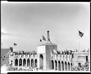 Arched entryway to the Los Angeles Memorial Coliseum with the Olympic flame and flags flying, 1932