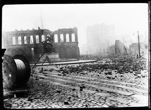 Earthquake damage in an unidentified street, showing a statue of men pulling the fulcrum of a large machine