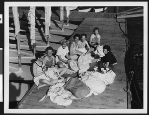 Seven women in sports attire seated on a dinghy dock sewing sails