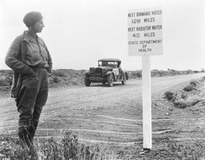 Female motorist in the desert standing next to a warning sign for next available water, ca.1920-1930