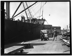 Dockworkers pushing a cart stacked with cargo next to a ship in Los Angeles Harbor