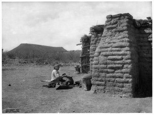 Pima Indian woman sitting on the ground making pottery by an adobe and grass hut, Gila River Reservation