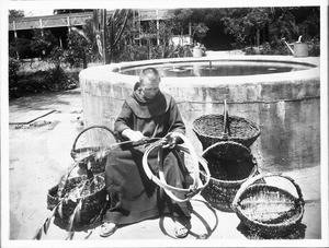 Monk sitting making baskets near the fountain in the garden at Mission Santa Barbara, ca.1898-1901