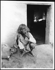 Hopi Indian man crouching outside and lighting a cigarette, ca.1900