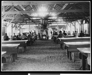 People eating in the interior of an open-air dining pavilion, showing a dirt floor, ca.1900