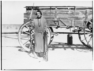 Old woman holding rifles and revolvers in front of a wagon