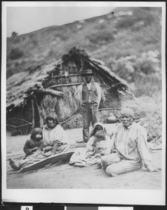 Group of Indian men, women, and children sitting in front of a thatched dwelling