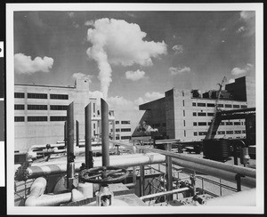 Exterior view of large unidentified factory, showing steam pipes in foreground, ca.1950