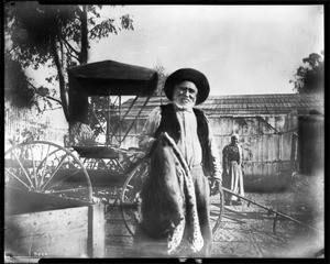 Portrait of Fecundo Reyes holding a horse-blanket, standing to the side of a shanty barn