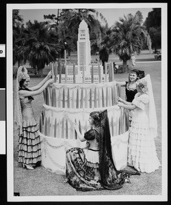 Young women in Spanish costumes placing candles cake that has a model of Los Angeles City Hall on top, ca.1930