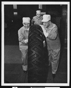 Three workers rolling a large tire, ca.1950