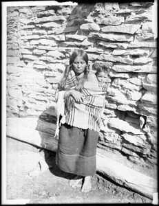 Hopi Indian mother carrying her baby on her back outside a native stone dwelling, ca.1900