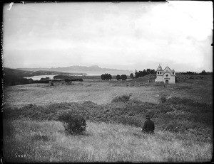 Man sitting in a grassy field looking toward Mission Carmel (San Carlos Borromeo de Carmelo) in Monterey, California, ca.1890-1897