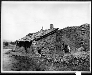 Old woman standing outside her adobe indian house at the Mission San Fernando, ca.1898