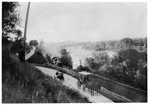 Two horse-drawn vehicles travel next to a train, showing a river below