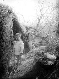 Apache Indian boy standing in front of a "Kan" or brush house, ca.1900