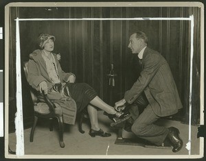 Man helping a woman try on shoes in a store, ca.1920