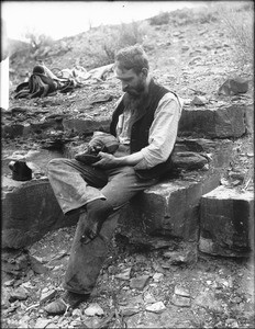 George Wharton James examining his worn-out shoe at River Camp on Bass Trail, Grand Canyon, ca.1900-1930