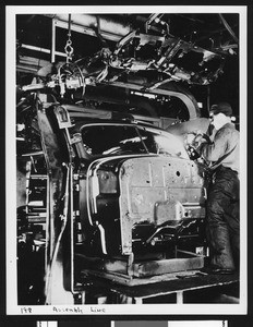 Interior view of an automobile factory showing a worker on an assembly line, ca.1925