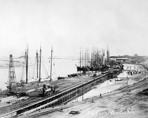 View of the Southern Pacific slip at San Pedro Harbor, showing a steam locomotive and oil derrick at left, ca.1900