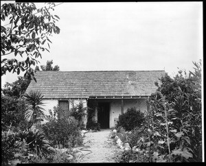 Exterior view of the Casa de Los Rios adobe, seen through a lush garden, ca.1890