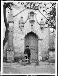 Father Grogan standing at the side door to the church of Mission San Buenaventura, California, ca.1904