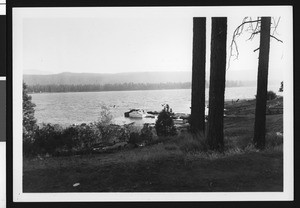 Big Bear Lake, showing trees and hills on opposite shore as well as the dock with boats at the near shore, ca.1950